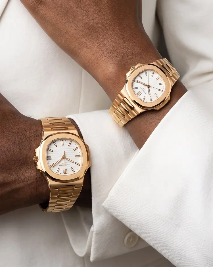 Two gold watches with white dials worn on crossed wrists, against the cuffs of a white suit